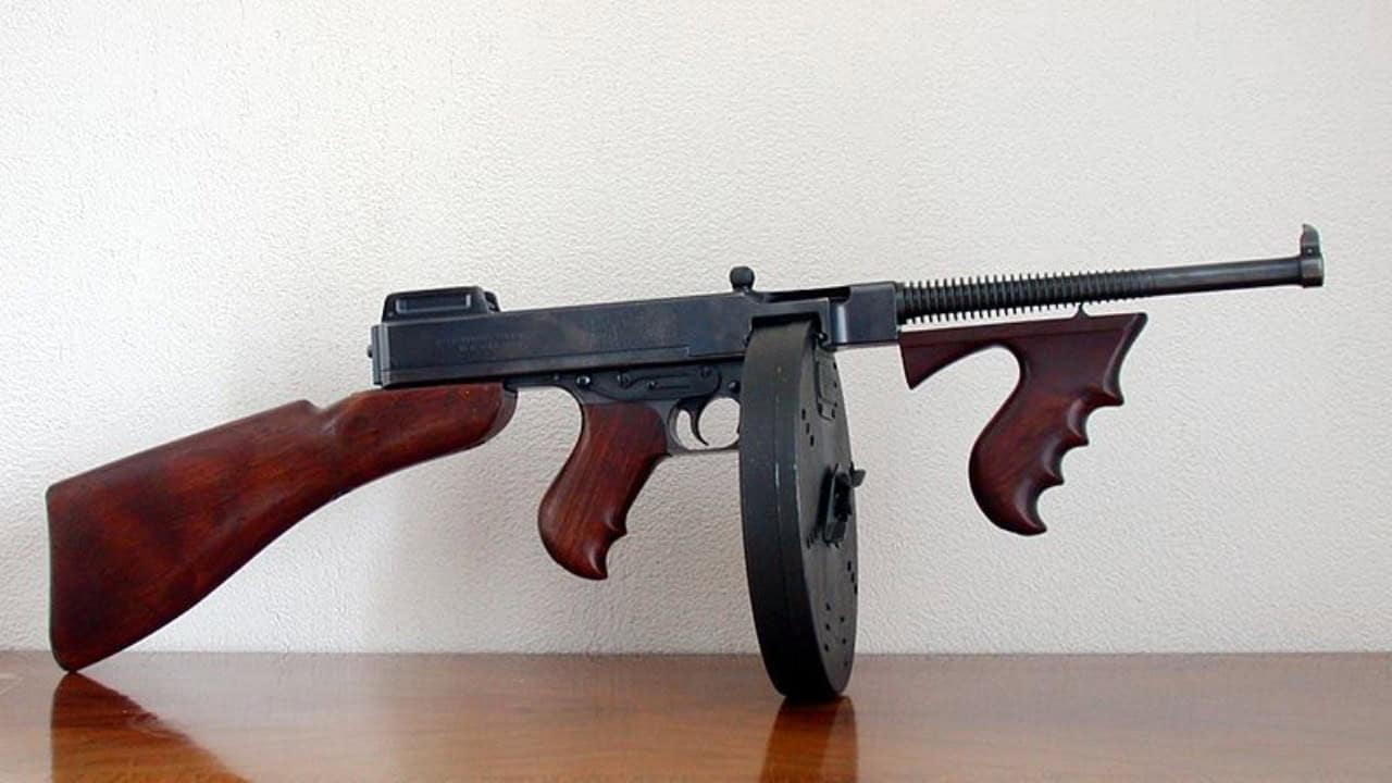 A Thompson submachine gun with a wooden stock and grip, and a round drum magazine, placed against a light-colored wall—a truly iconic weapon.
